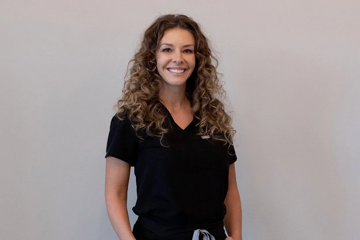 Photo of Brittney from Liv Aesthetics. She stands in front of a softly lit background, wearing a white coat and smiling confidently, representing professionalism and a welcoming presence at the med spa.