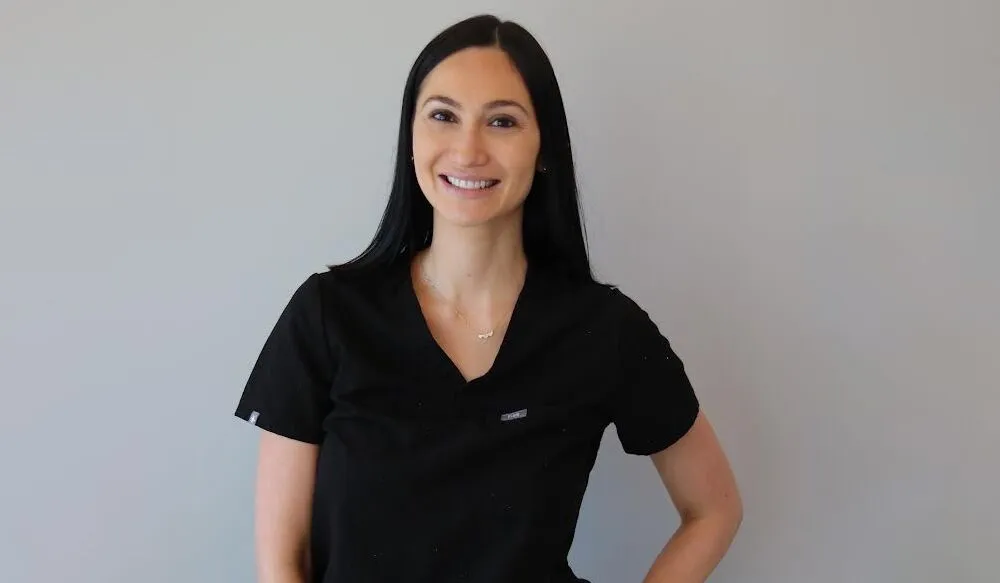Stacey from Liv Aesthetics LLC smiles warmly while wearing a black uniform, posed against a clean, softly lit background. Her approachable expression conveys confidence and professionalism.