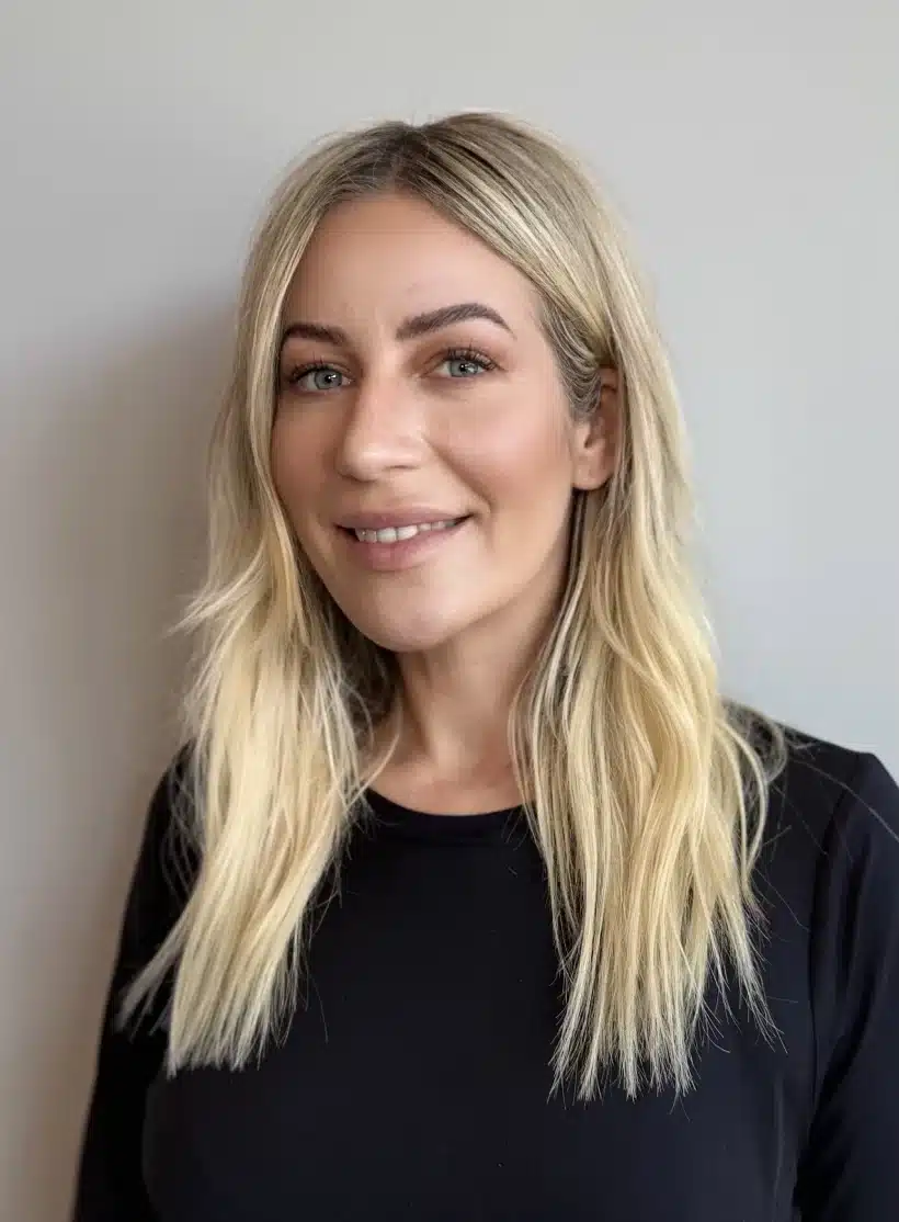 A smiling esthetician Kate wearing a black uniform, stands in front of a white wall with framed artwork, exuding a friendly and professional demeanor.