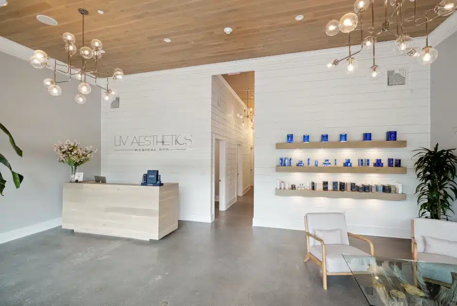 Sleek and inviting medical spa lobby featuring a wood reception desk, floating product display shelves, modern lounge chairs, and decorative lighting under a wood-paneled ceiling. A large plant and fresh flowers add natural warmth to the clean, minimalist design.