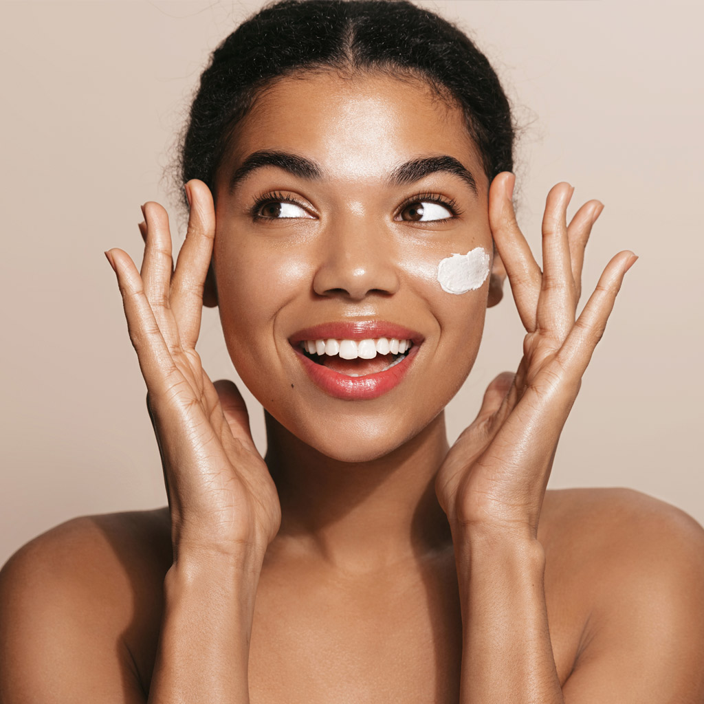 Happy smiling woman applies facial moisturizing cream, nourish her skin, stands over brown background
